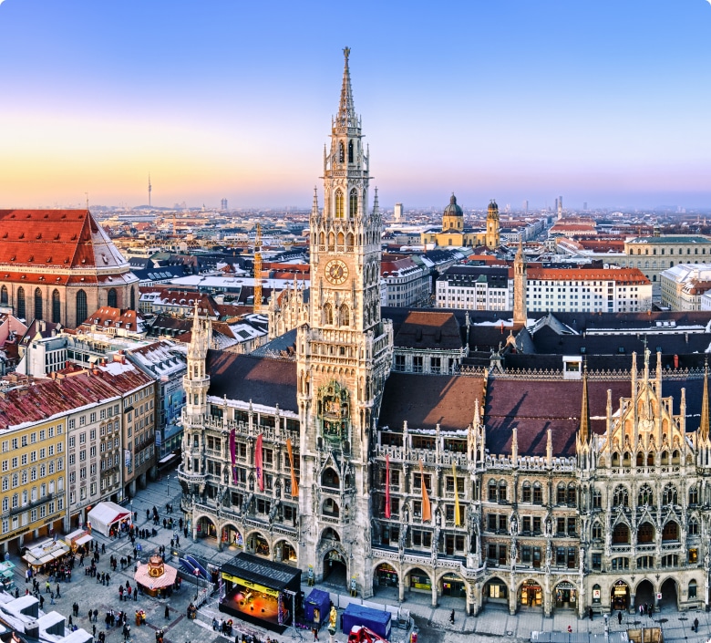 Luftaufnahme von München bei Sonnenuntergang. Hervorzuheben ist das gotische Neue Rathaus mit seinen Türmen, umgeben von Gebäuden und Straßen.